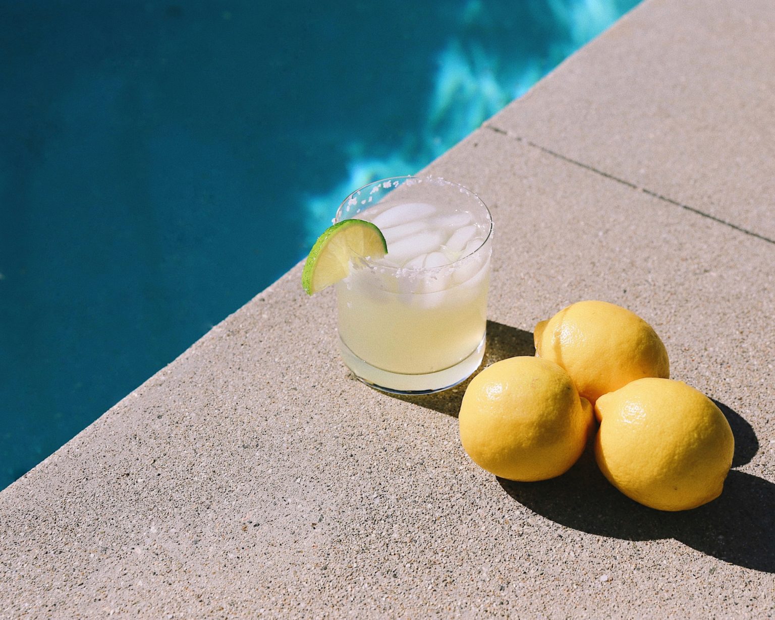 Glass of lemonade with lime garnish beside fresh lemons by a pool