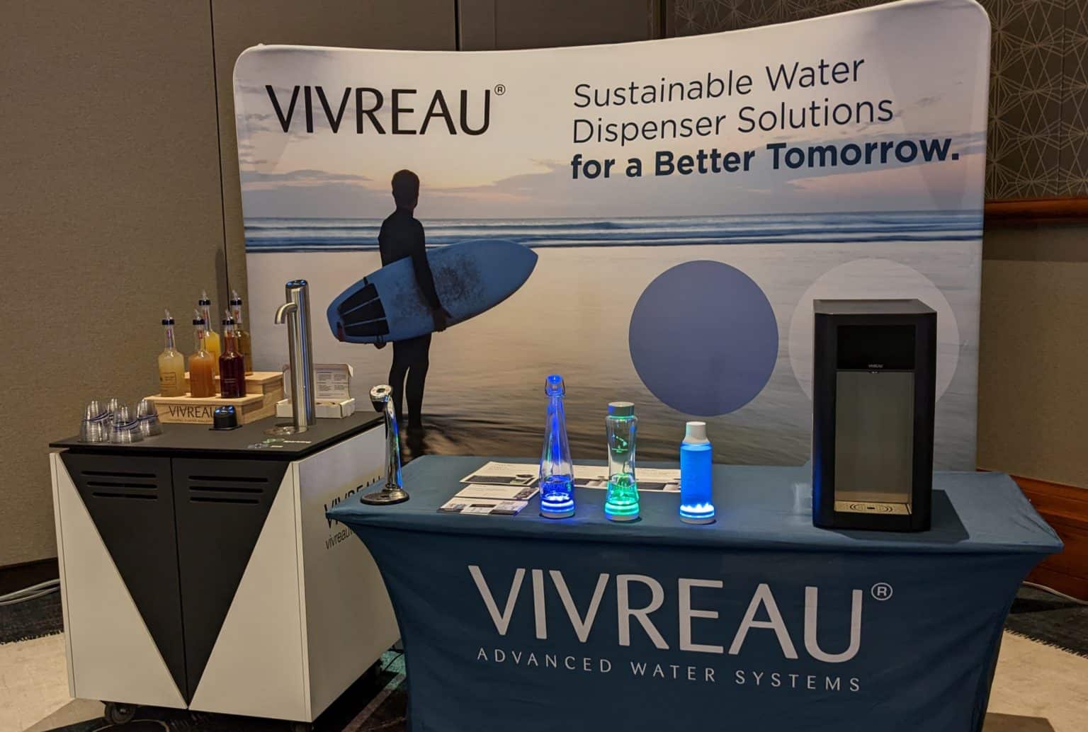 Vivreau booth showcasing sustainable water dispenser solutions with a backdrop featuring a surfer at sunset, a variety of water bottles, and beverage dispensers