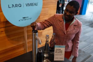 A woman using a Vivreau water dispenser at TED2023, with branded Vivreau and LARQ glass bottles nearby promoting sustainability