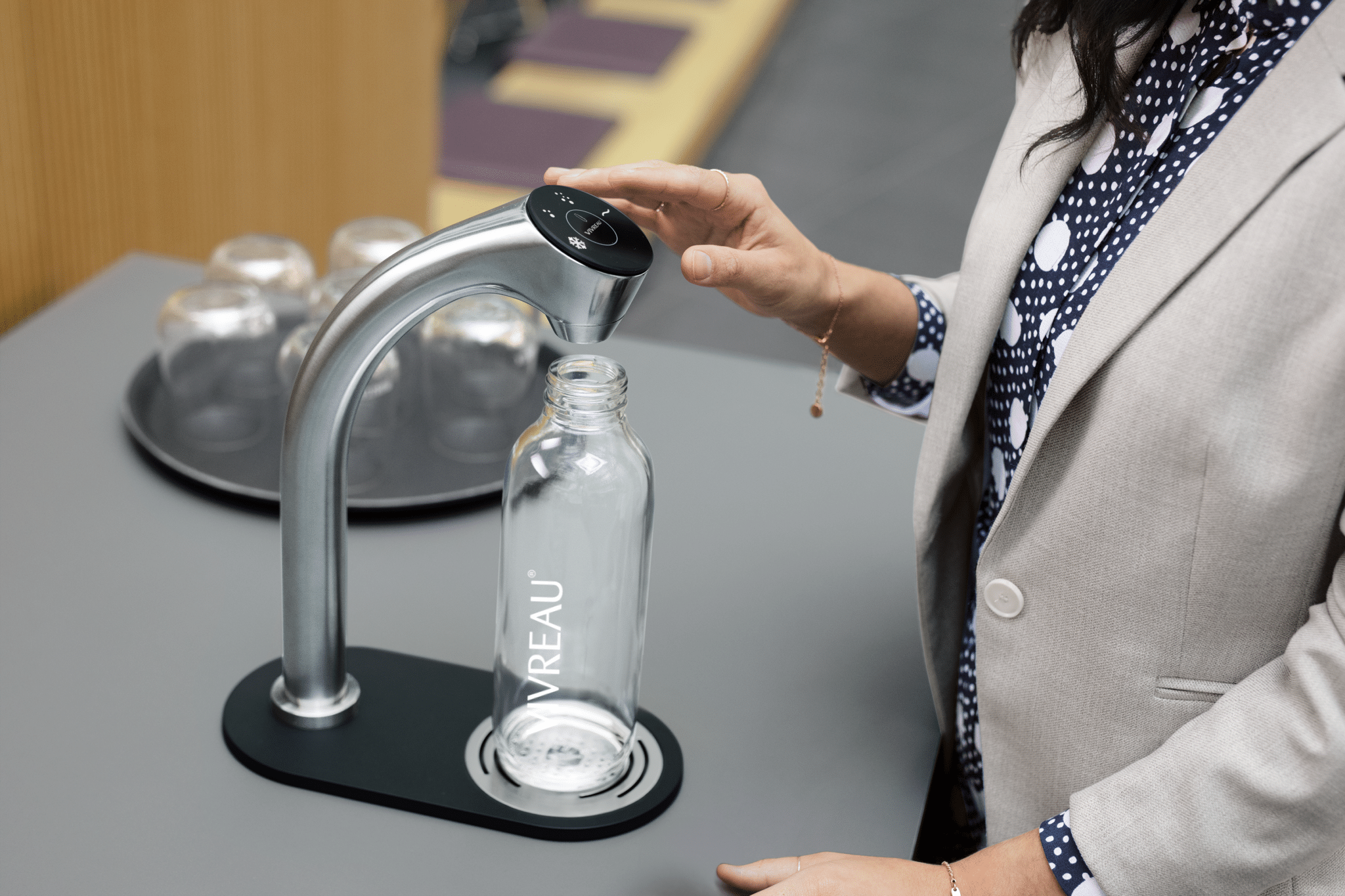 A person in a business suit refilling a reusable Vivreau glass water bottle from a modern touch-operated water dispenser