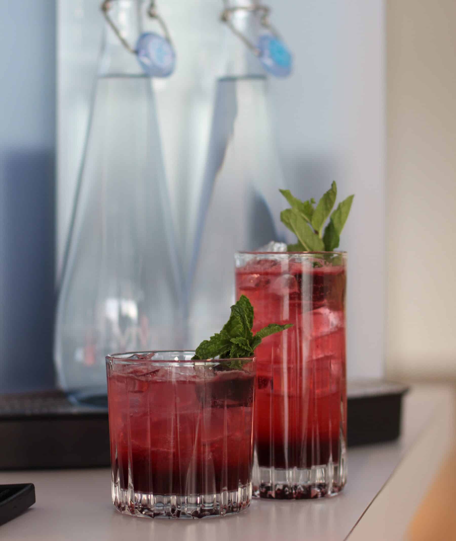 two glasses of spiced pomme mocktail garnished with mint leaves placed in front of glass water bottles