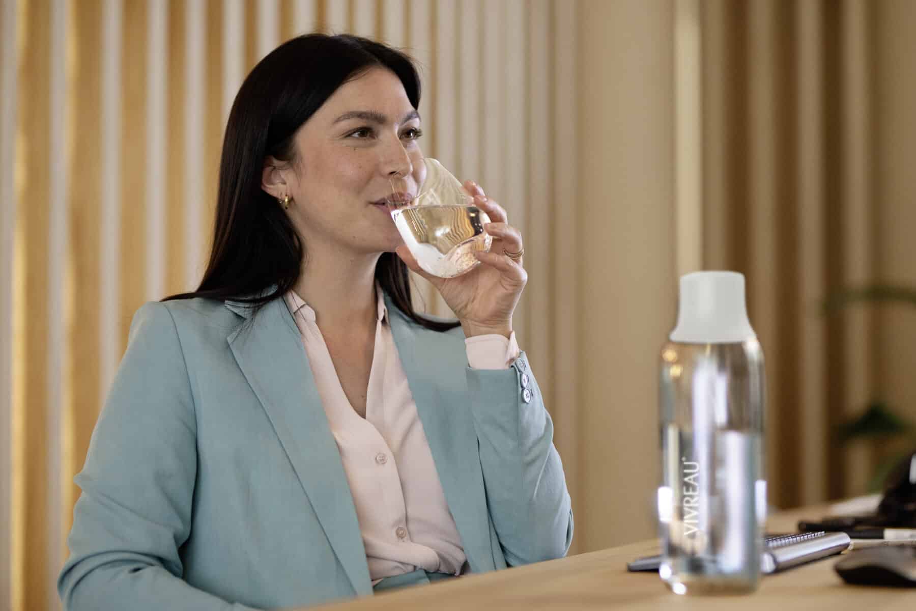 Woman in a light blue blazer drinking water from a glass with a Vivreau bottle on the desk