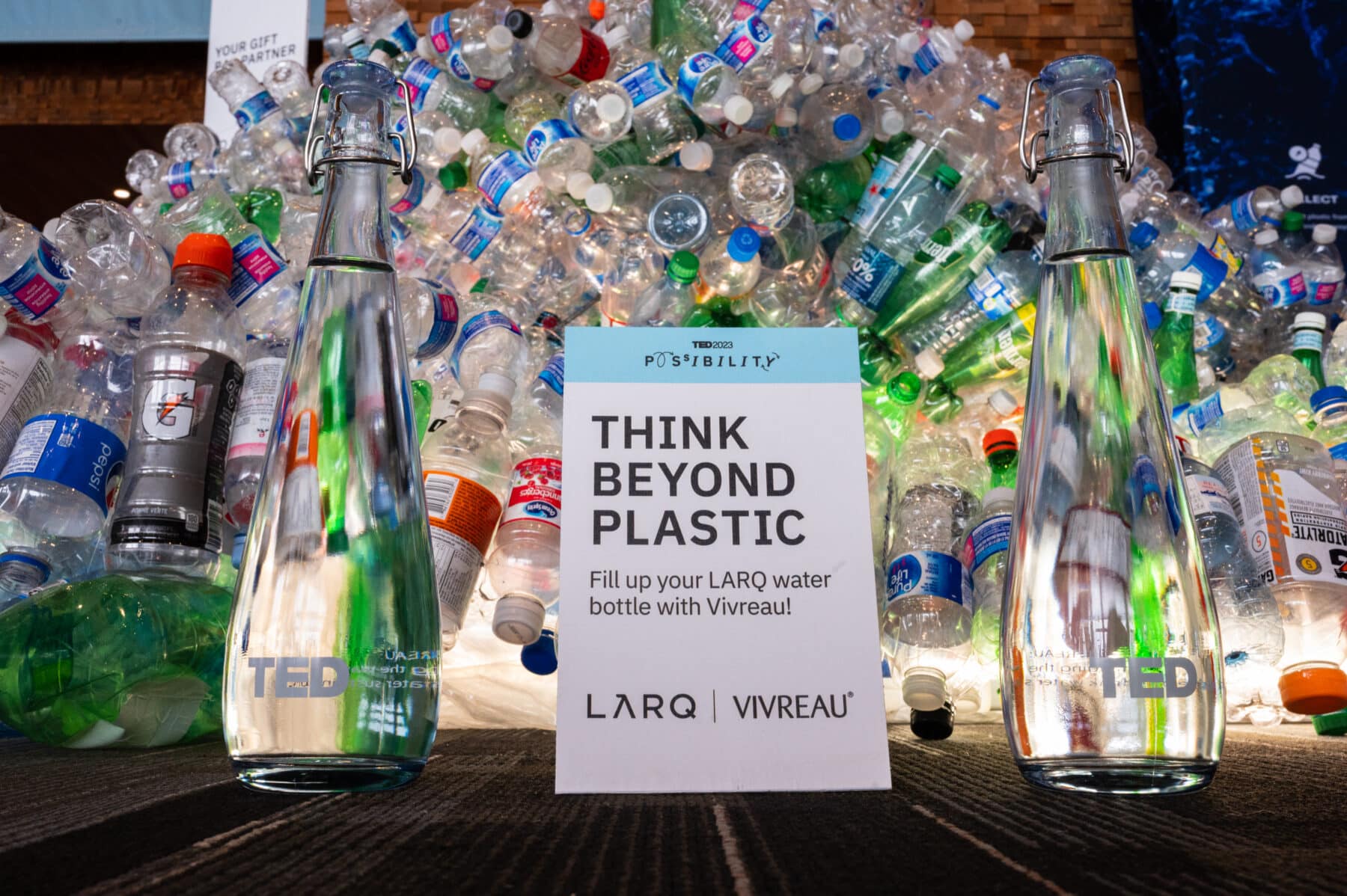 Glass water bottles branded with TED2023 and Vivreau placed in front of a display of plastic waste with a sign promoting sustainability and refillable bottles