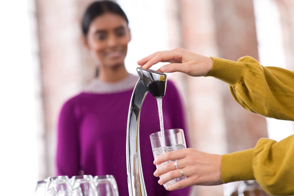 Person filling a glass with water from a Vivreau Vi Tap while another person observes in the background