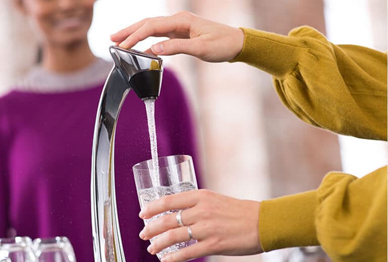 Hand filling a glass of water from a Vivreau dispenser with another person in the background